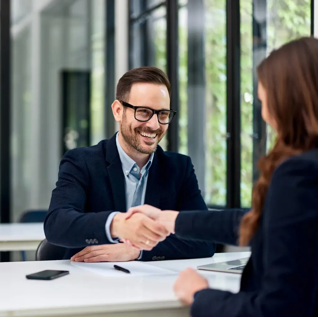 A man smiling while he shakes a woman's hand at a desk.